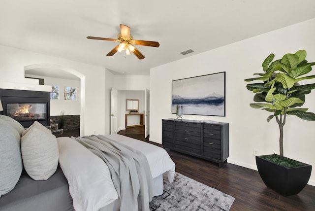 bedroom featuring visible vents, a glass covered fireplace, dark wood-style floors, baseboards, and ceiling fan