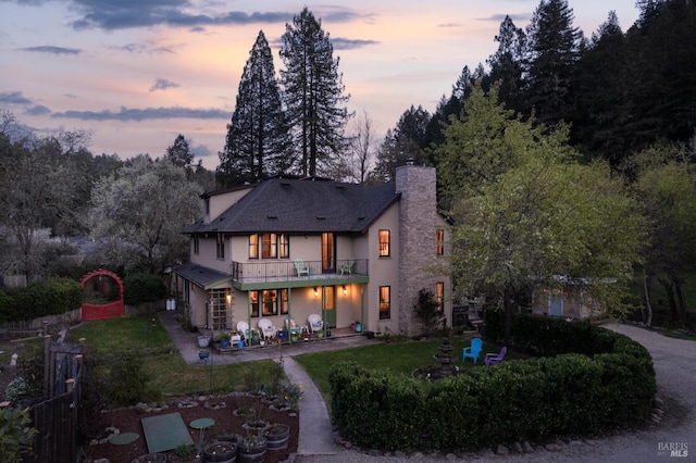 back of house at dusk featuring stucco siding, a lawn, a patio, a balcony, and a chimney