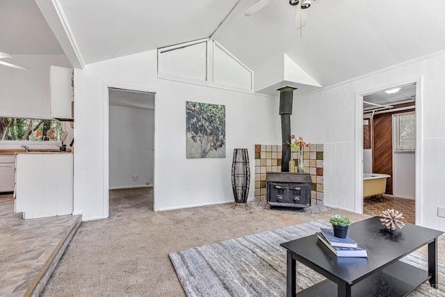 living area with ceiling fan, light colored carpet, a wood stove, high vaulted ceiling, and baseboards