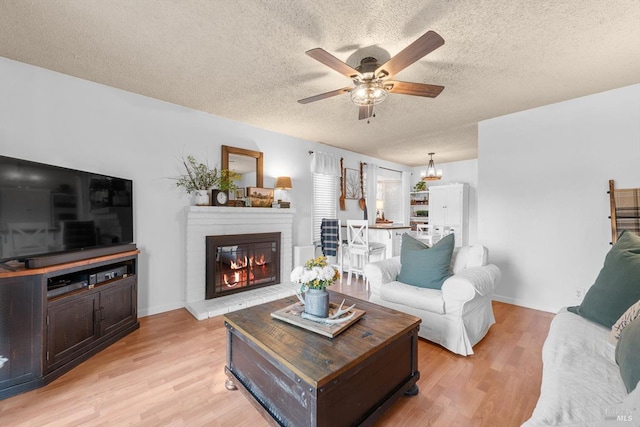 living room with a brick fireplace, ceiling fan with notable chandelier, light wood finished floors, and a textured ceiling