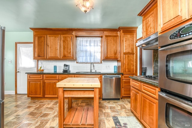 kitchen featuring tasteful backsplash, under cabinet range hood, brown cabinets, appliances with stainless steel finishes, and a sink