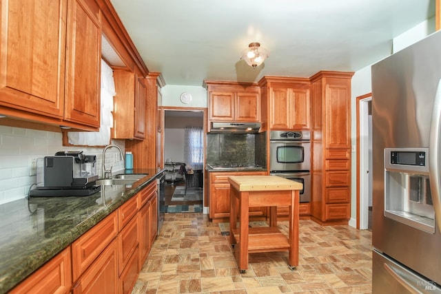 kitchen featuring a sink, stone finish flooring, under cabinet range hood, appliances with stainless steel finishes, and decorative backsplash