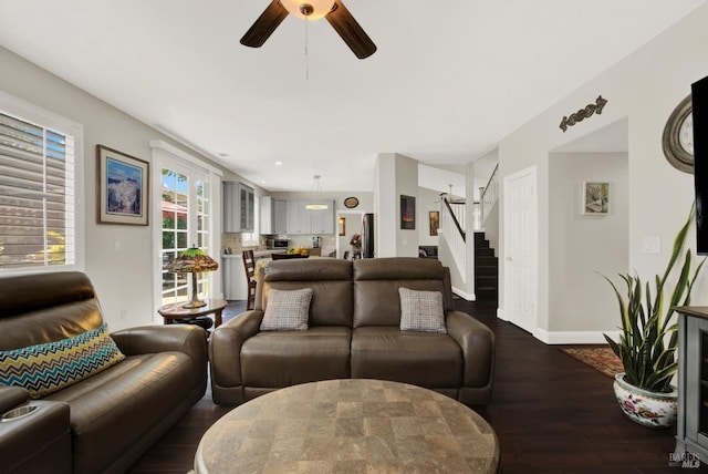 living area featuring baseboards, a ceiling fan, dark wood-style floors, and stairs