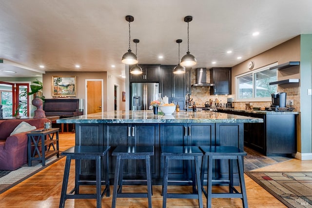 kitchen with a kitchen bar, stone counters, stainless steel refrigerator with ice dispenser, a large island, and wall chimney exhaust hood