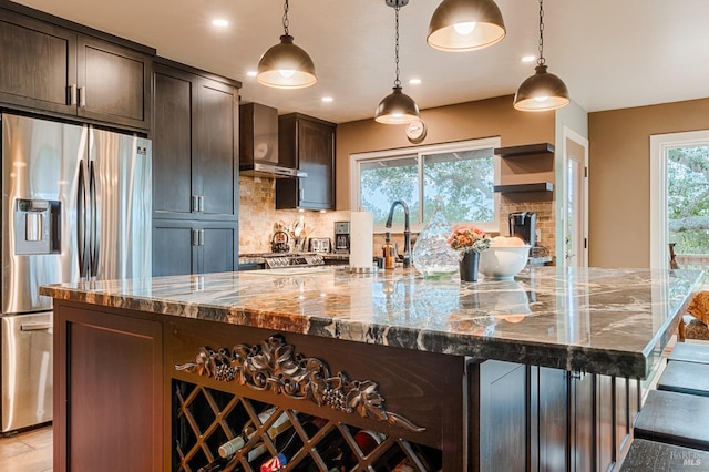 kitchen with tasteful backsplash, dark brown cabinets, dark stone counters, stainless steel refrigerator with ice dispenser, and wall chimney exhaust hood