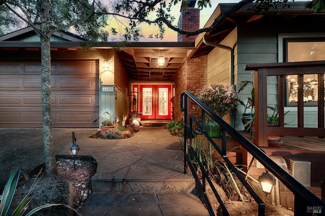 exterior entry at dusk with a chimney, french doors, an attached garage, and brick siding