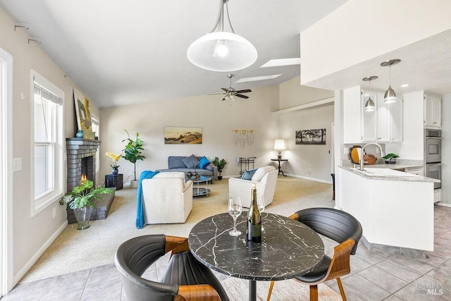 dining space featuring a brick fireplace, vaulted ceiling, light carpet, light tile patterned flooring, and a ceiling fan