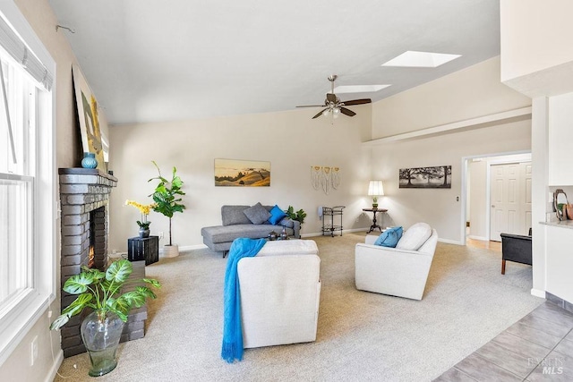 living room with light carpet, a ceiling fan, vaulted ceiling with skylight, baseboards, and a brick fireplace