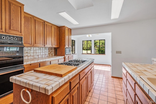 kitchen featuring tile counters, decorative backsplash, brown cabinets, light tile patterned flooring, and stainless steel appliances