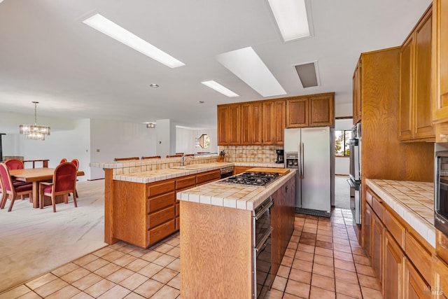 kitchen featuring tile countertops, a peninsula, a sink, stainless steel appliances, and a center island