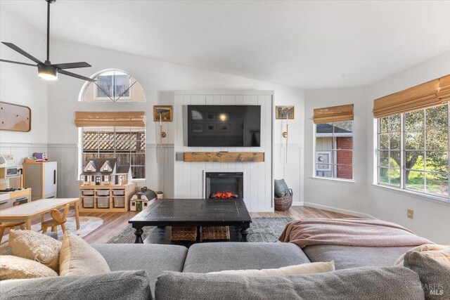 living area with a lit fireplace, ceiling fan, wainscoting, and wood finished floors