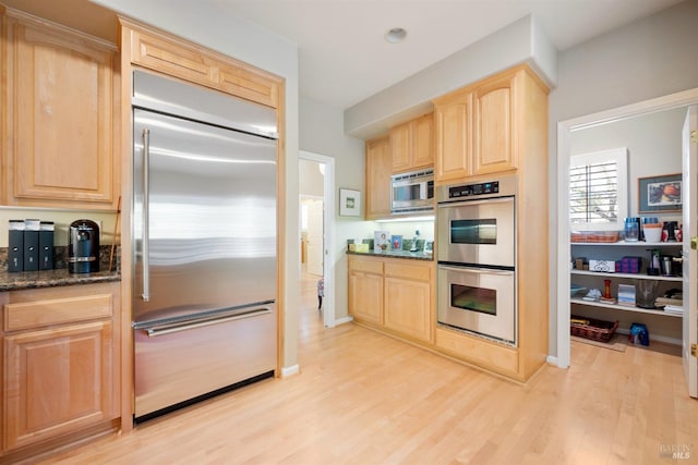 kitchen featuring light brown cabinetry, built in appliances, light wood-style flooring, and dark stone countertops