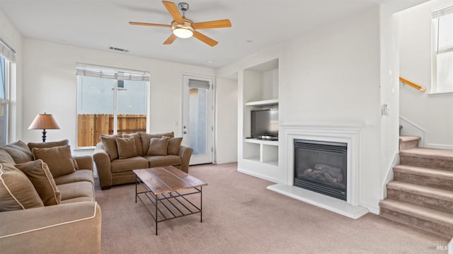 living room with built in shelves, light carpet, a ceiling fan, a glass covered fireplace, and stairway