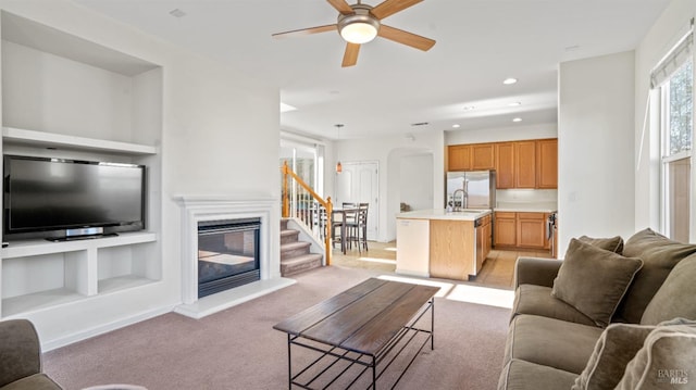 living room featuring built in shelves, ceiling fan, light carpet, arched walkways, and a glass covered fireplace