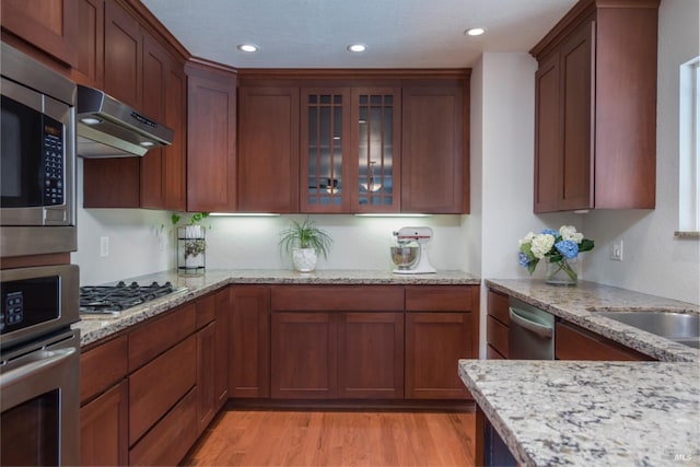 kitchen with under cabinet range hood, stainless steel appliances, light stone countertops, and light wood-style flooring