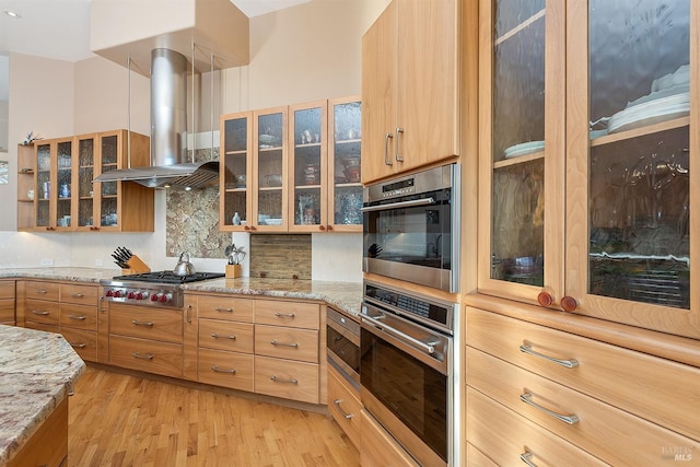 kitchen with light wood finished floors, stainless steel appliances, glass insert cabinets, wall chimney range hood, and tasteful backsplash