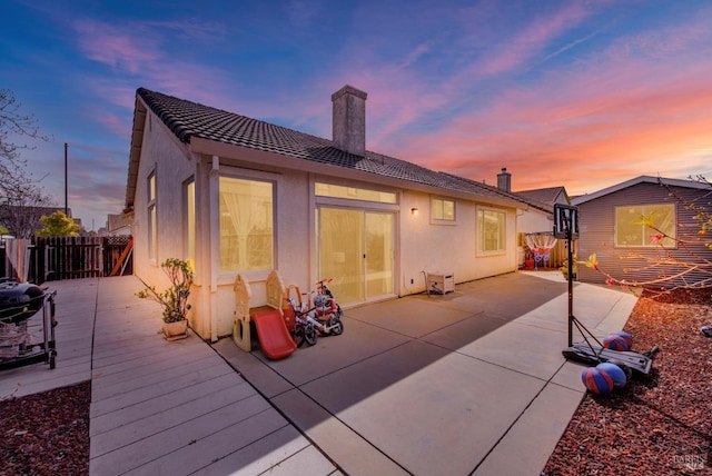 back of house at dusk with a chimney, a deck, fence, a patio, and stucco siding