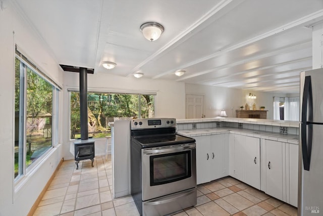 kitchen featuring beam ceiling, white cabinetry, tile countertops, stainless steel appliances, and a wood stove