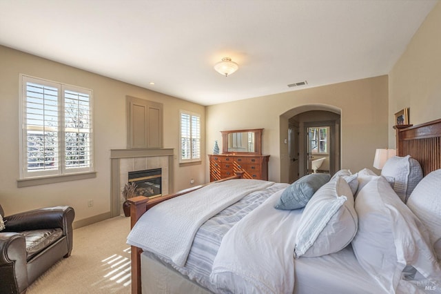 bedroom with light colored carpet, visible vents, a tiled fireplace, and arched walkways