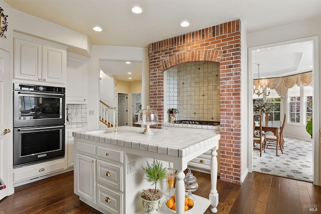kitchen featuring double oven, white cabinets, a sink, tile counters, and dark wood-type flooring