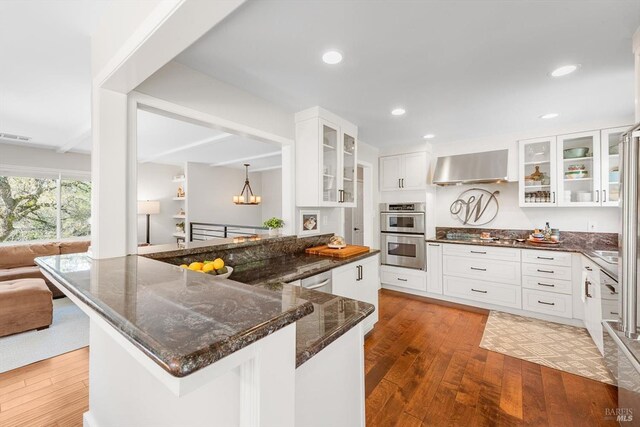 kitchen with dark stone countertops, wall chimney range hood, wood-type flooring, and stainless steel double oven