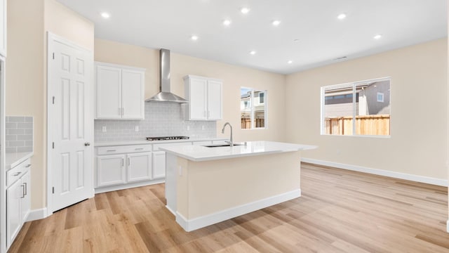 kitchen featuring gas cooktop, wall chimney range hood, a center island with sink, a sink, and light wood-style flooring