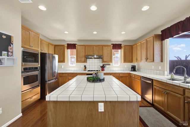 kitchen featuring a sink, tile counters, under cabinet range hood, appliances with stainless steel finishes, and a center island