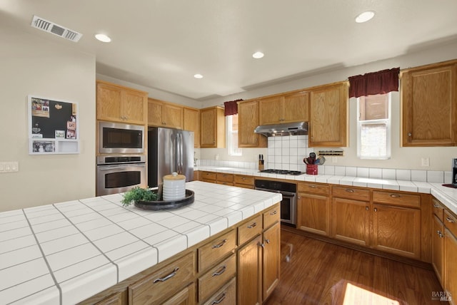 kitchen featuring visible vents, under cabinet range hood, tile countertops, stainless steel appliances, and dark wood-style flooring