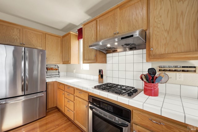 kitchen featuring light wood-style flooring, under cabinet range hood, tasteful backsplash, appliances with stainless steel finishes, and tile counters