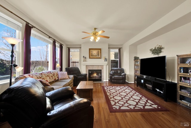 living area with a ceiling fan, a tiled fireplace, wood finished floors, and baseboards