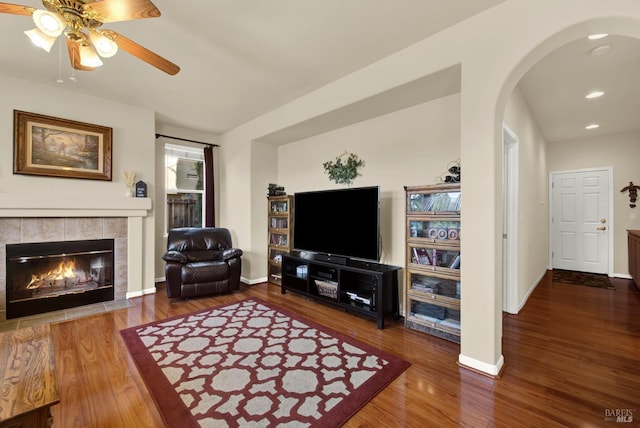 living area featuring baseboards, a tiled fireplace, wood finished floors, arched walkways, and a ceiling fan
