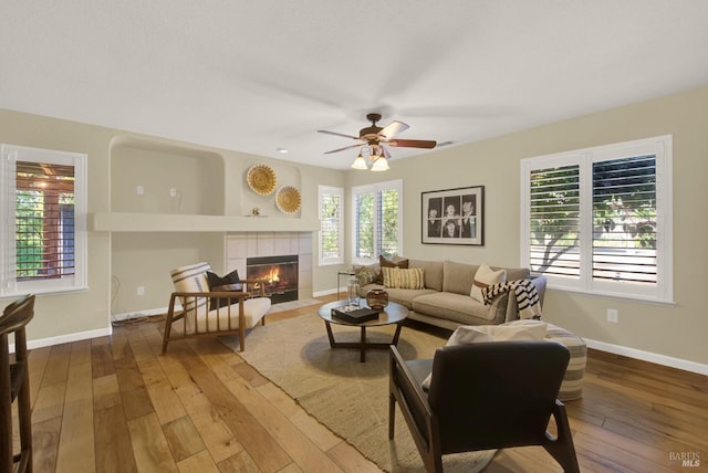 living room with baseboards, a tiled fireplace, a ceiling fan, and wood-type flooring