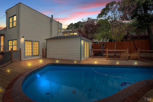 pool at dusk with fence, an outdoor pool, a deck, and an outbuilding
