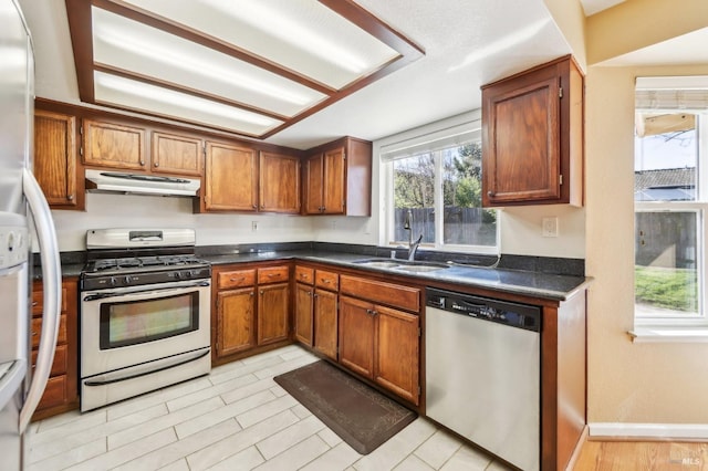 kitchen with stainless steel appliances, dark countertops, under cabinet range hood, a sink, and brown cabinetry