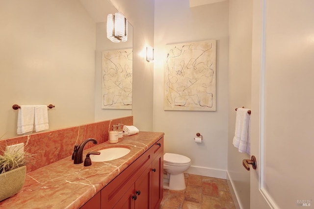 bathroom featuring toilet, baseboards, stone finish flooring, vanity, and decorative backsplash