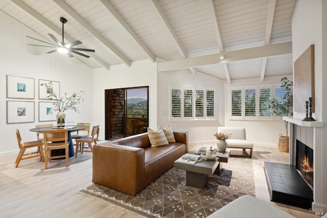 living room with beam ceiling, wood finished floors, a warm lit fireplace, ceiling fan, and wood ceiling