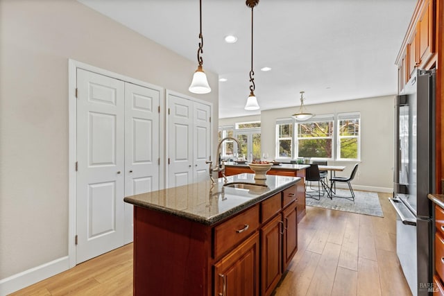 kitchen with dark stone countertops, an island with sink, a sink, light wood-style floors, and freestanding refrigerator