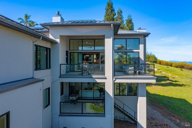 rear view of property featuring a standing seam roof, a chimney, stucco siding, a balcony, and metal roof