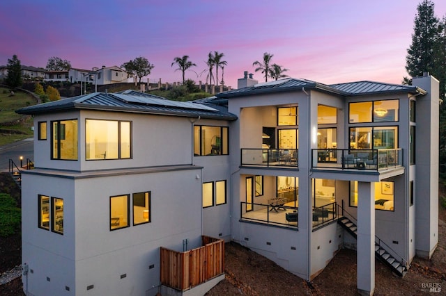 back of property at dusk featuring a standing seam roof, metal roof, a balcony, solar panels, and a chimney