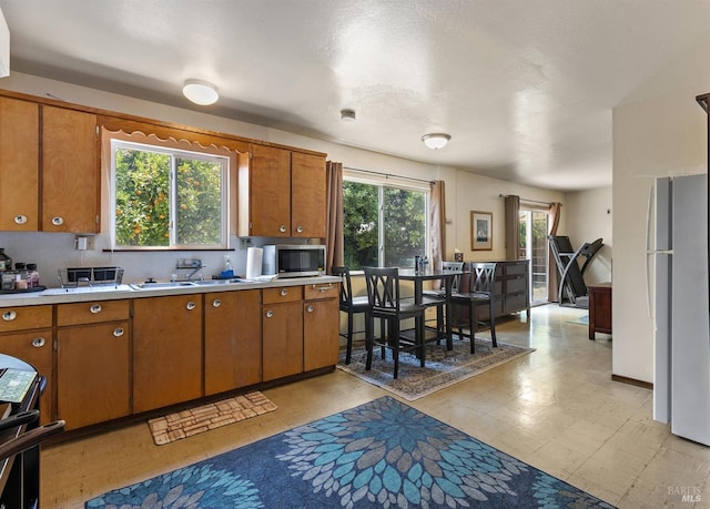 kitchen with stainless steel microwave, light countertops, freestanding refrigerator, brown cabinets, and a sink