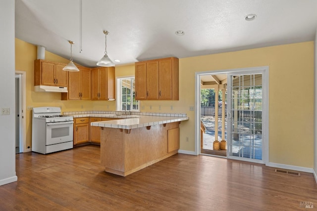 kitchen with white range with gas cooktop, a peninsula, under cabinet range hood, dark wood-type flooring, and tile countertops