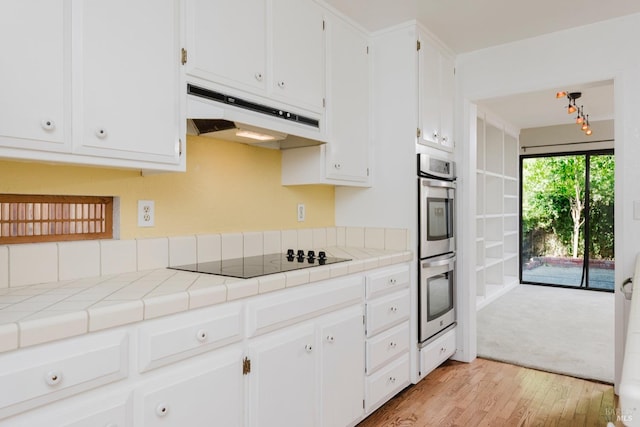kitchen featuring light wood-style floors, tile countertops, white cabinetry, under cabinet range hood, and black electric cooktop