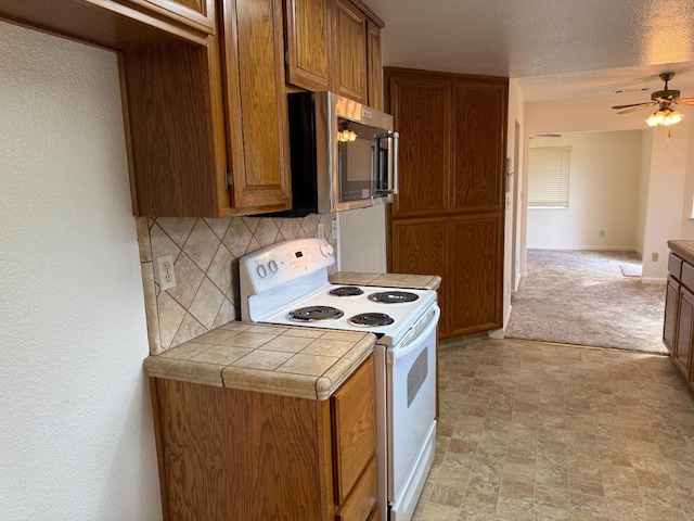 kitchen with white range with electric stovetop, tile counters, brown cabinetry, stainless steel microwave, and decorative backsplash