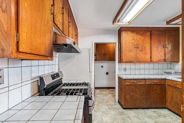 kitchen with stainless steel gas stove, tile counters, under cabinet range hood, brown cabinets, and backsplash