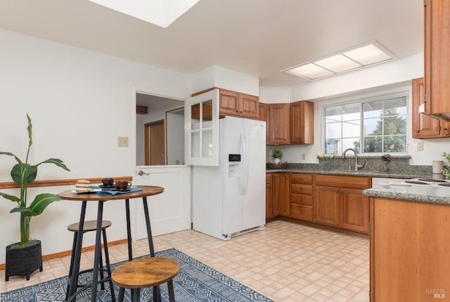 kitchen with white refrigerator with ice dispenser, dark stone counters, a sink, light floors, and brown cabinets