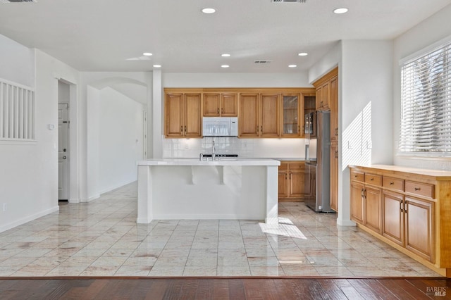 kitchen featuring light countertops, an island with sink, freestanding refrigerator, white microwave, and brown cabinets