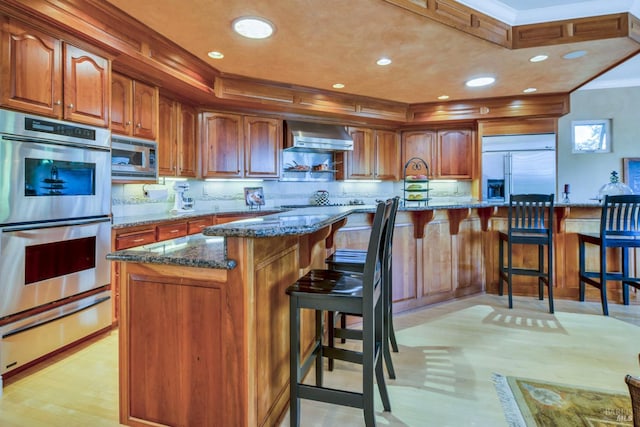 kitchen featuring wall chimney exhaust hood, dark stone counters, a kitchen bar, and built in appliances