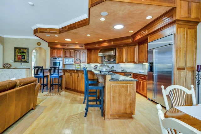 kitchen with crown molding, built in appliances, wall chimney exhaust hood, a kitchen breakfast bar, and arched walkways