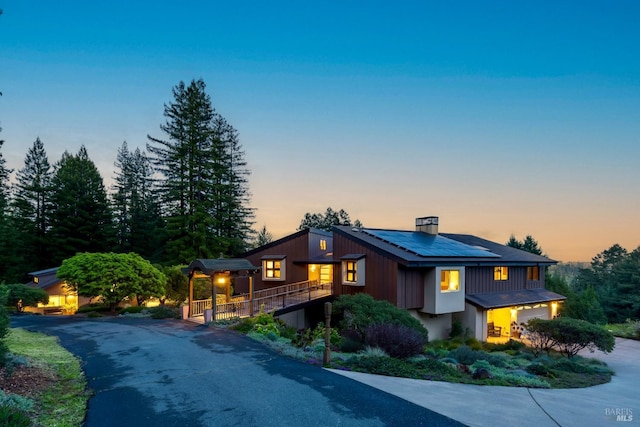 view of front of property with board and batten siding, roof mounted solar panels, a garage, a chimney, and driveway