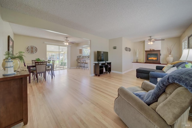 living area featuring a textured ceiling, light wood-type flooring, a glass covered fireplace, and a ceiling fan